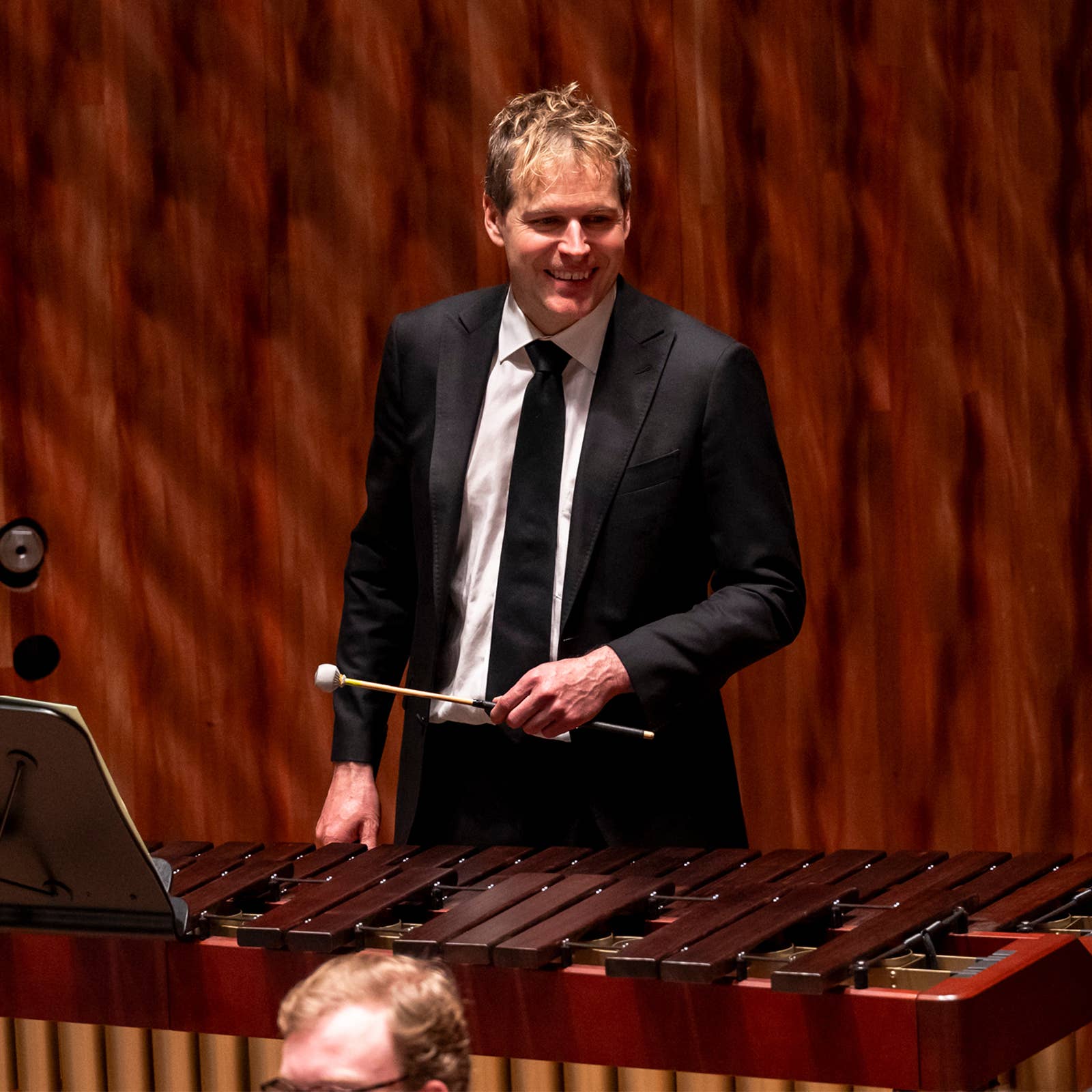 Man in a suit playing a marimba with a mallet, standing on a stage with a wooden backdrop.
