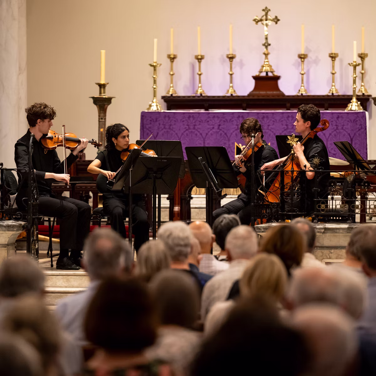 String quartet performs in a church, with musicians holding violins and a cello onstage behind music stands.