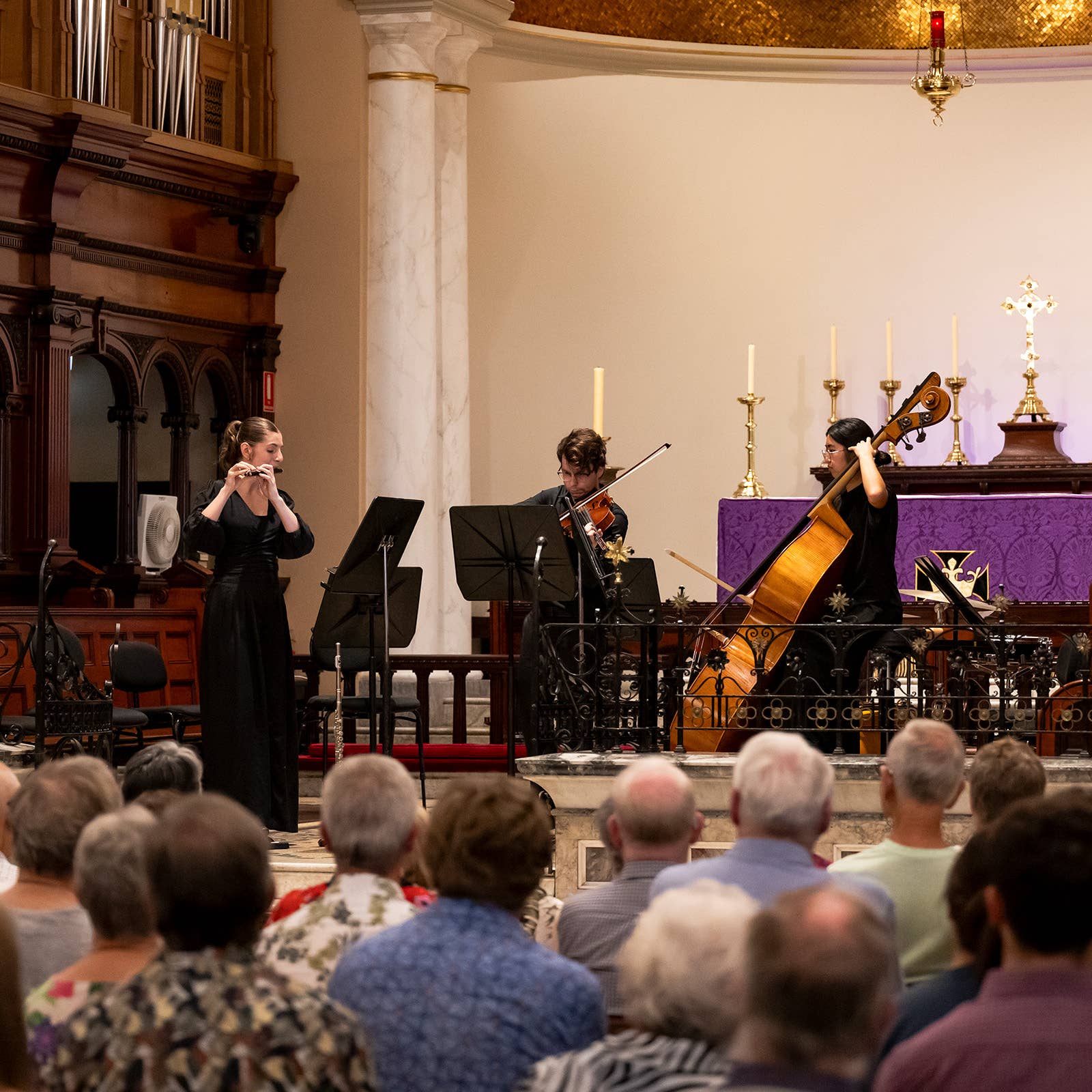 Musicians perform in a church during a concert, candles and purple altar cloth behind seated audience in foreground.