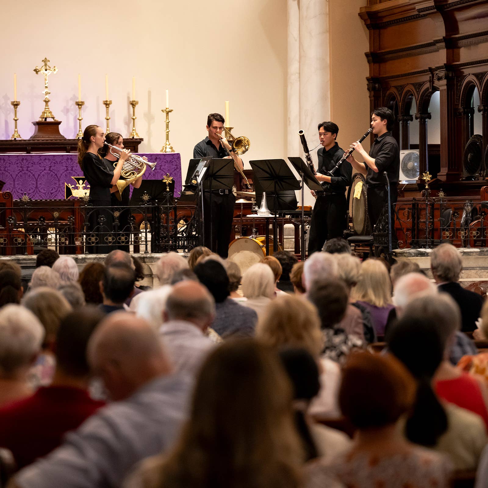 Musicians perform in a church, playing French horn, trombone, clarinet, and trumpet on stage.