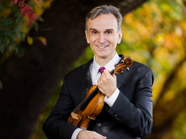 Smiling man in a suit holding a wooden violin outdoors with blurred autumn foliage in the background