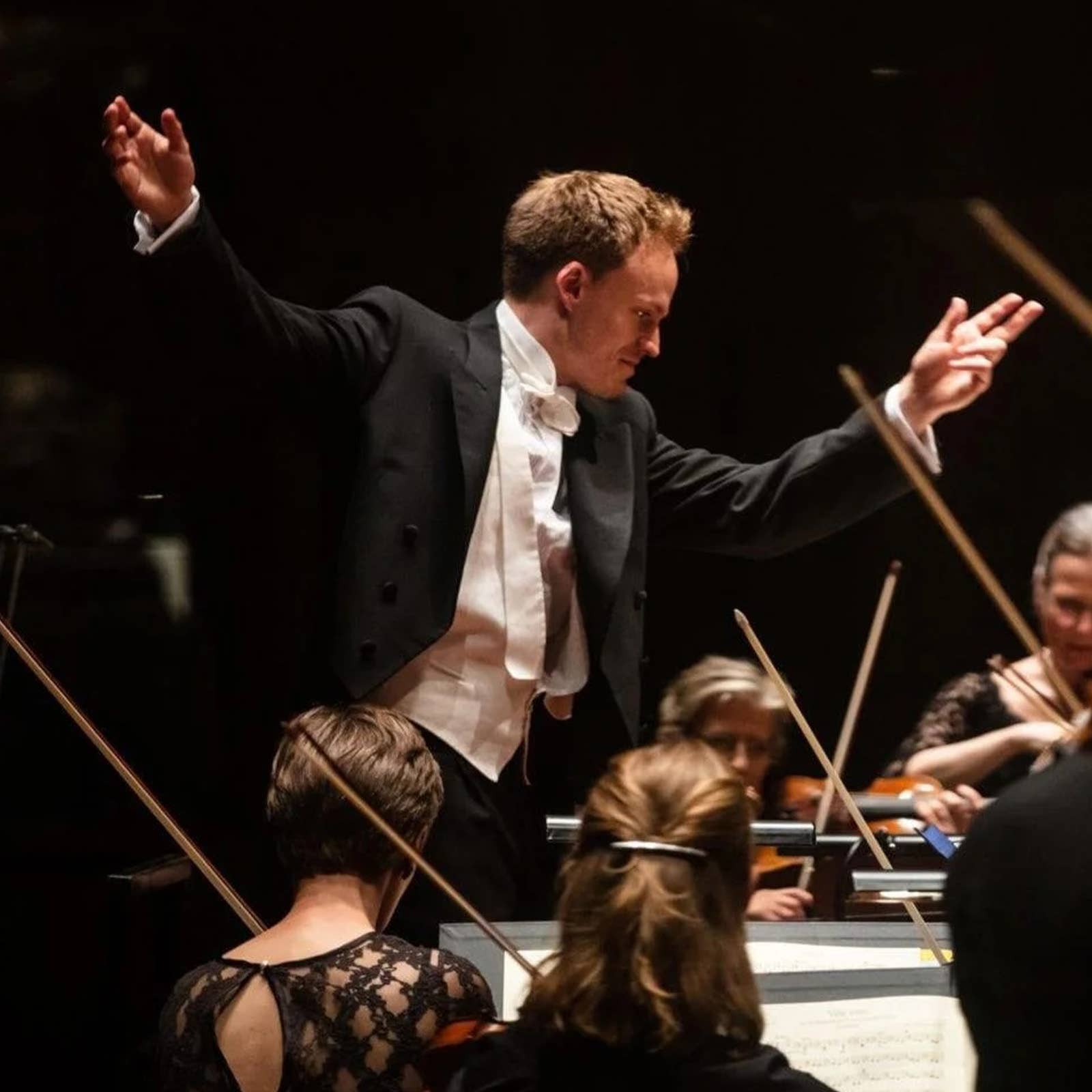 Conductor in a dark suit leads an orchestra with both hands raised, surrounded by musicians and string instruments.