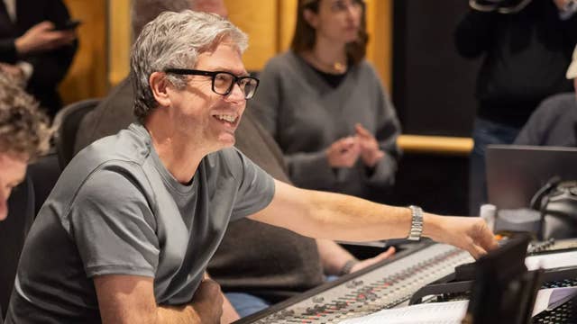 Man with gray hair and glasses smiles while working at a control console in a studio.