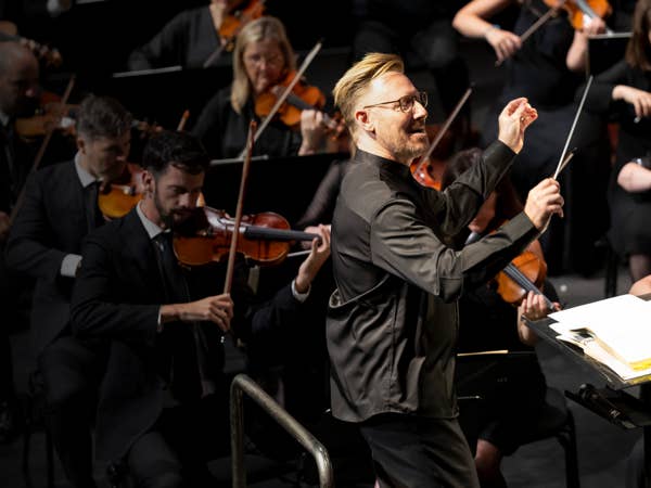 Conductor in a concert hall leads an orchestra as violinists play behind him under bright stage lights