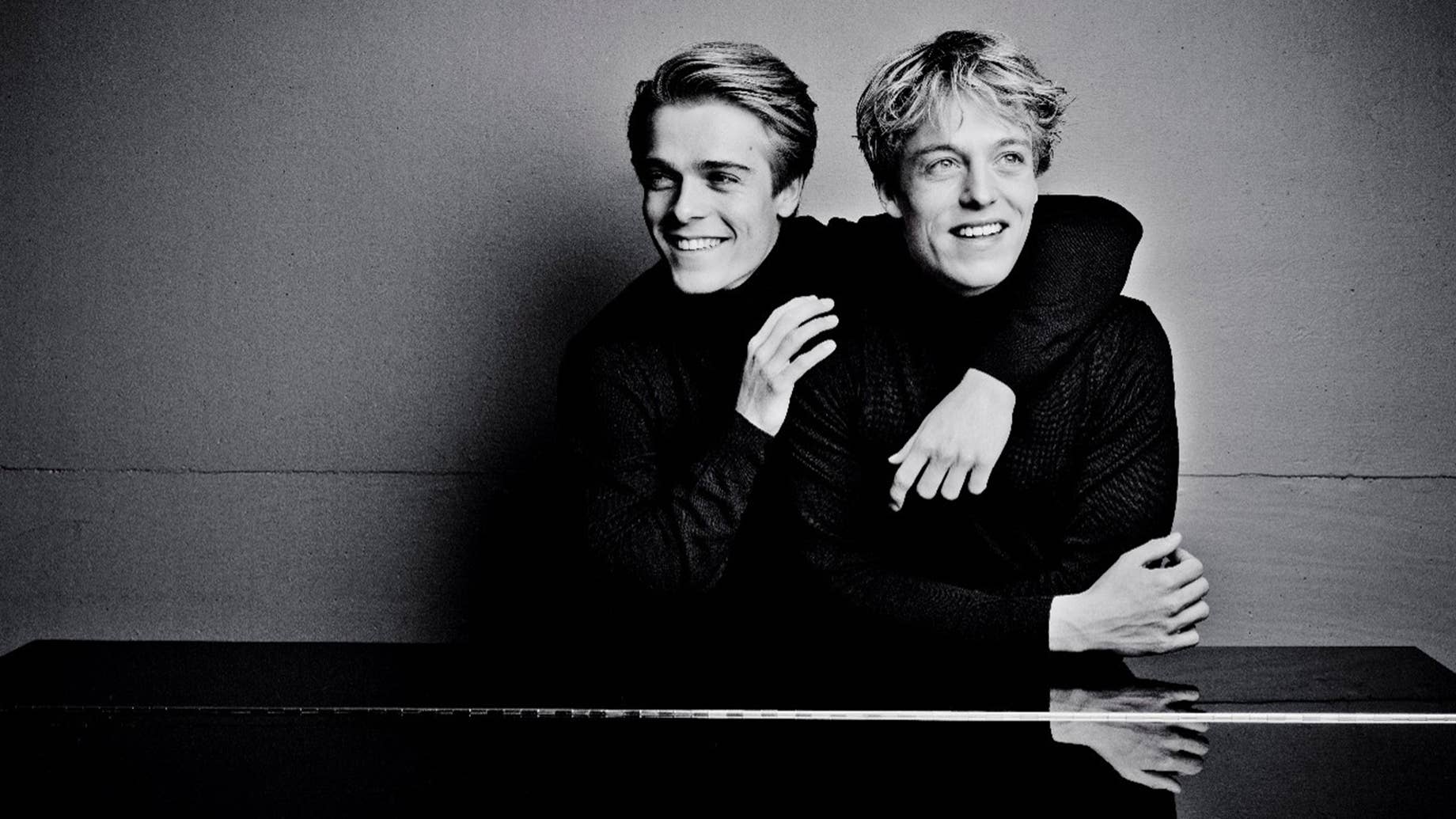 Two brothers sitting together smiling in a black-and-white portrait, one behind the other with arms resting forward and leaning on a piano