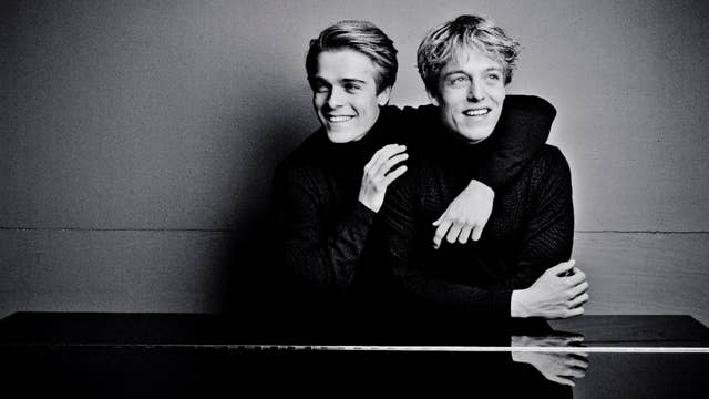 Two brothers sitting together smiling in a black-and-white portrait, one behind the other with arms resting forward and leaning on a piano