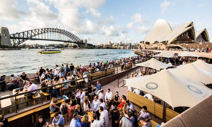 Crowded outdoor bar near Sydney Opera House with view of Sydney Harbour Bridge and ferry on the water under a partly cloudy sky.