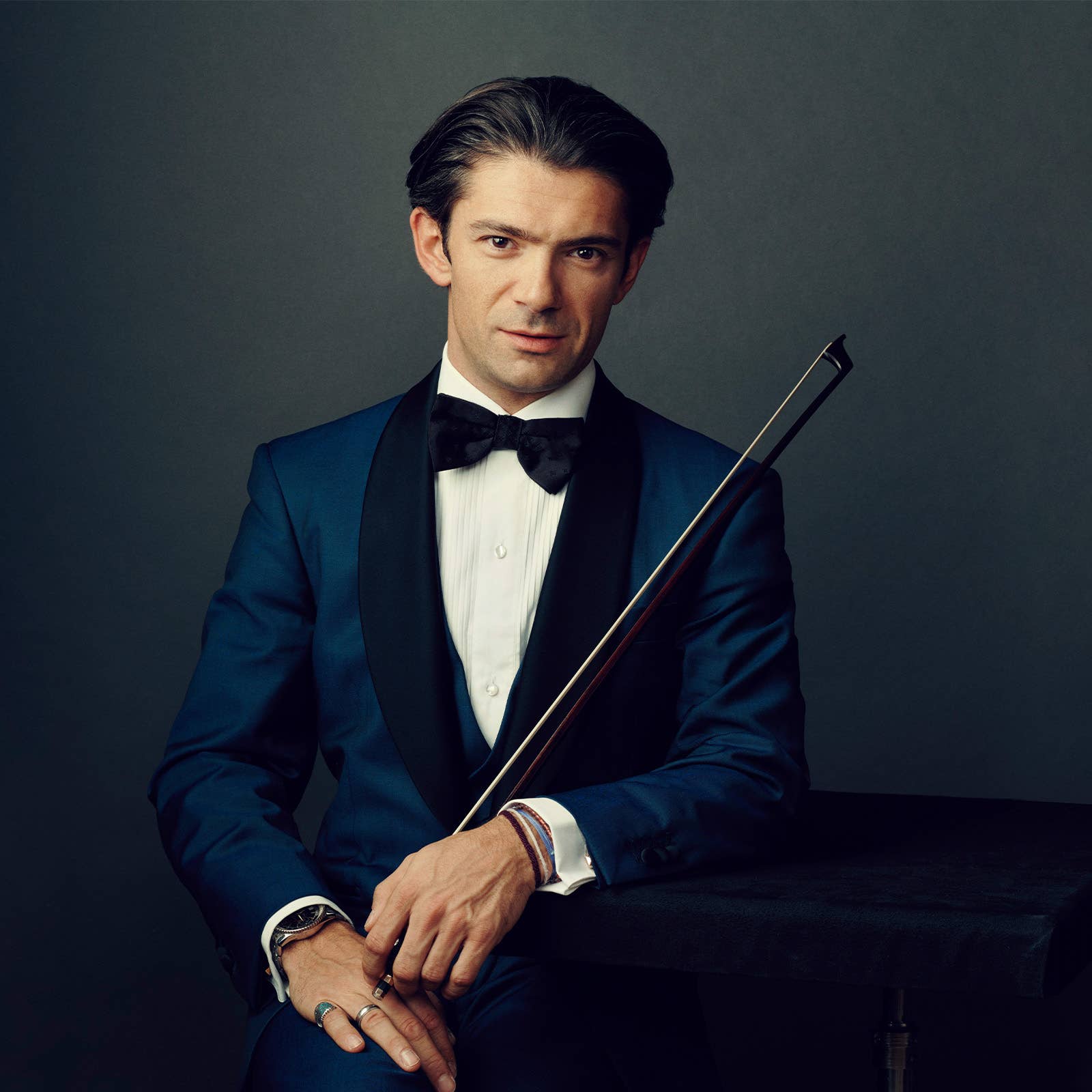 Smiling man in tuxedo holding an instrumental bow, posed against a dark studio background.