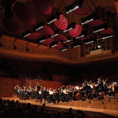 Orchestra performs on a concert hall stage under red spotlighted petals and stage lights overhead.