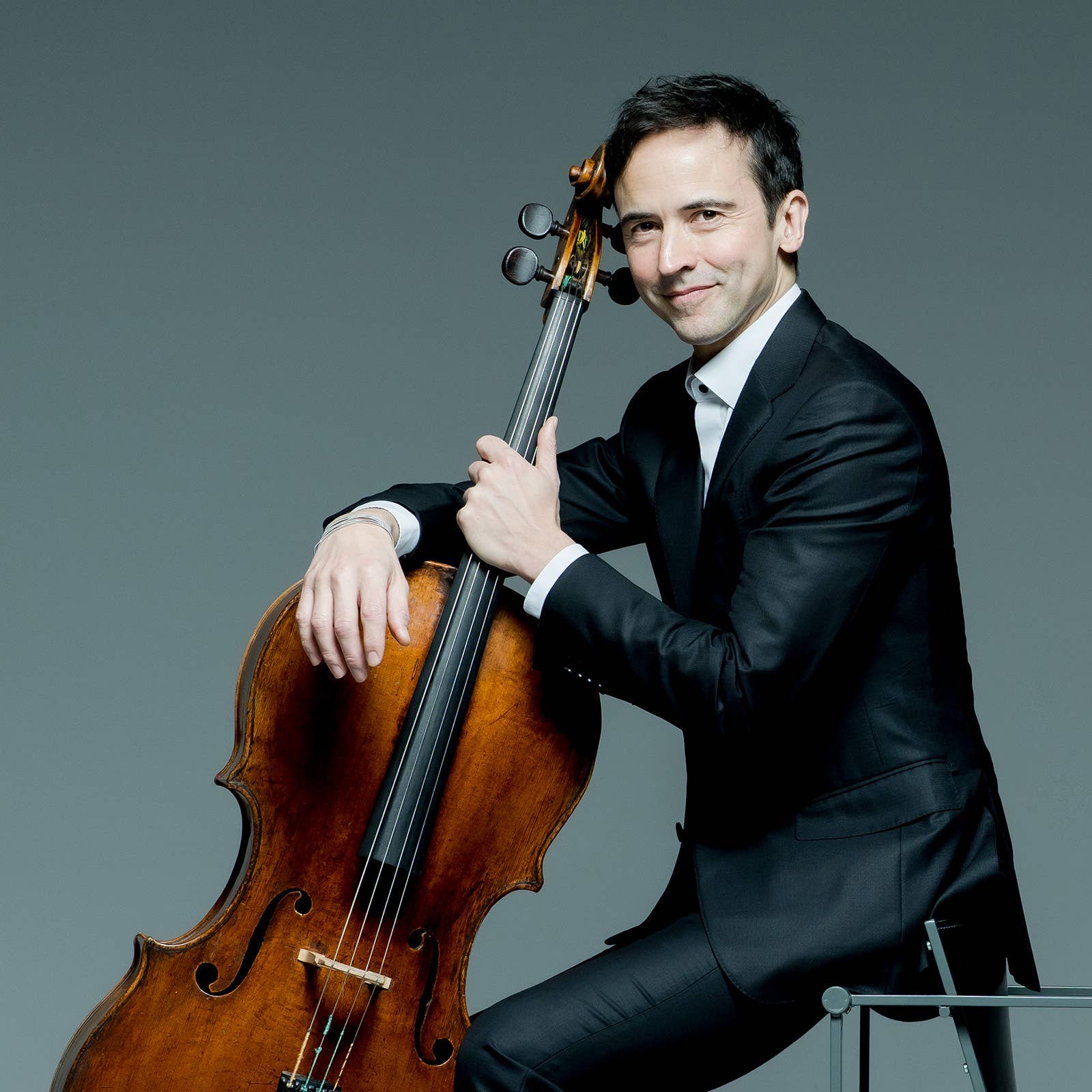 Man in a suit holding a cello, smiling with a bow against a gray background