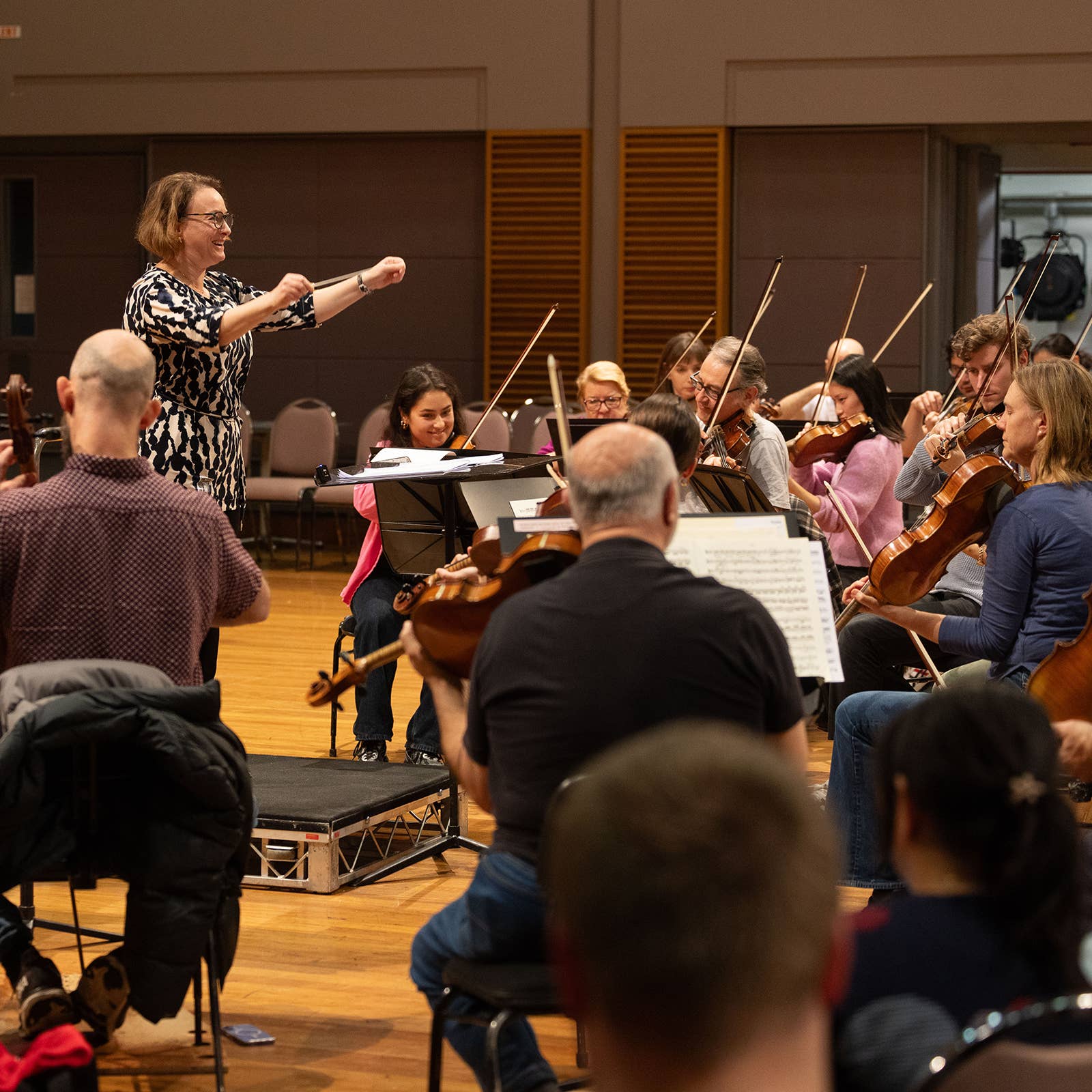 Conductor in patterned dress leads an orchestra rehearsal as musicians play violins and string instruments in a hall