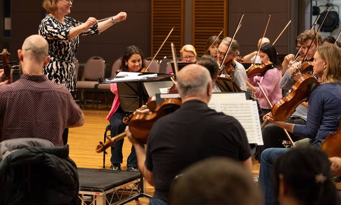 Conductor in patterned dress leads an orchestra rehearsal as musicians play violins and string instruments in a hall