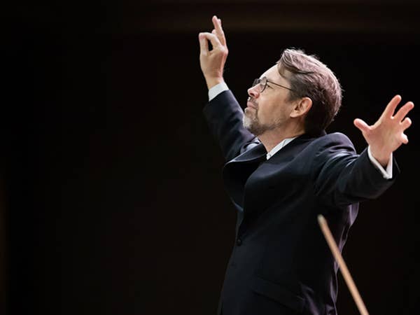 Man in suit with glasses raises both hands during performance on a dark stage.
