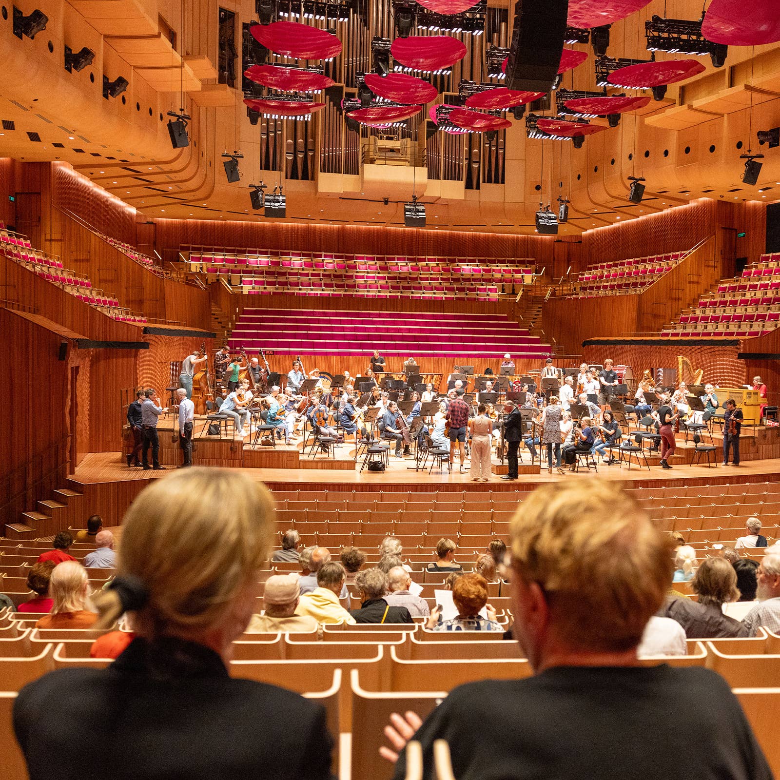 Orchestra rehearsing on stage in a concert hall with an audience seated in the foreground.