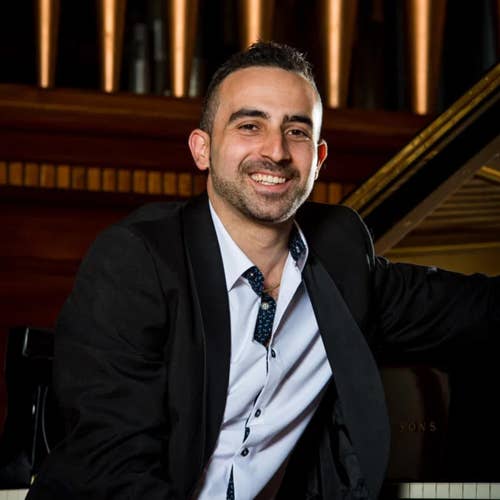 Smiling man in a dark suit and patterned tie seated in a warmly lit room with a piano behind him