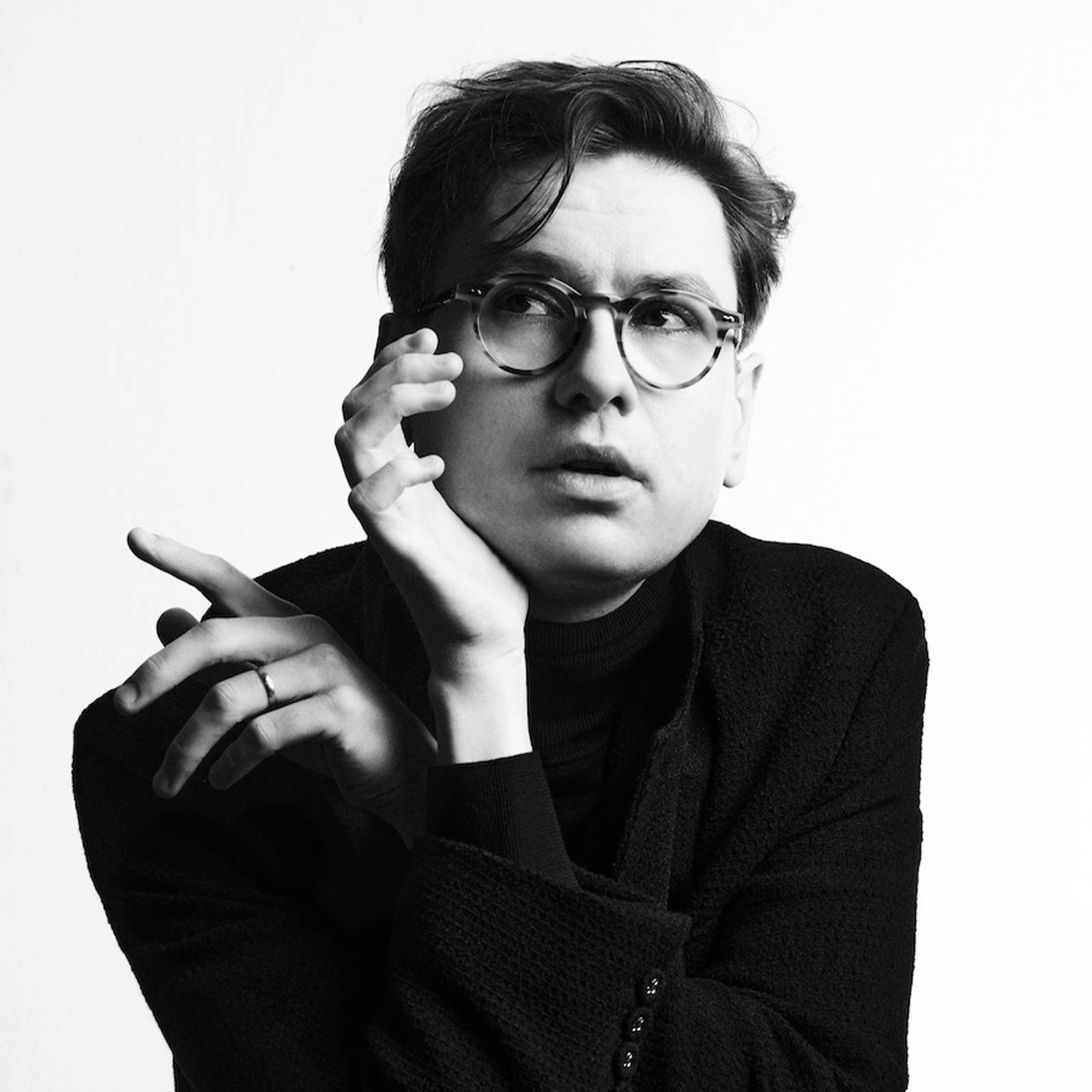Black-and-white portrait of a man with round glasses holding his chin and gesturing with a hand.