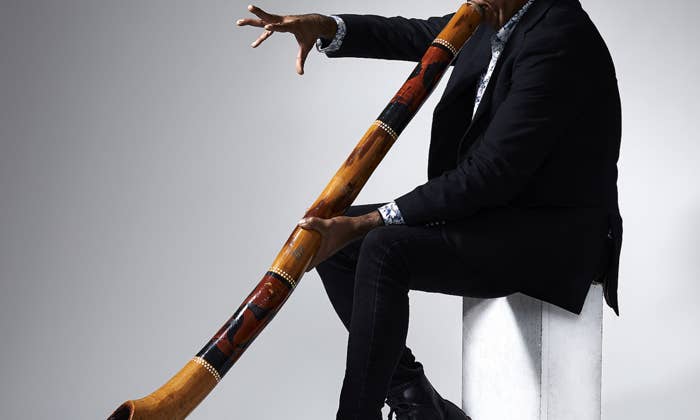 Man in a black suit playing a decorated didgeridoo while seated on a white block against a gray background.