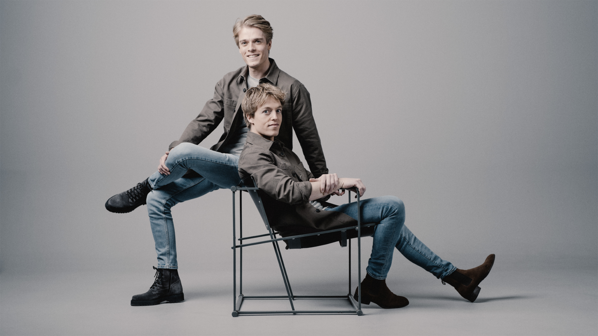 Two brothers in jackets and jeans pose together on a studio chair against a plain light gray background.