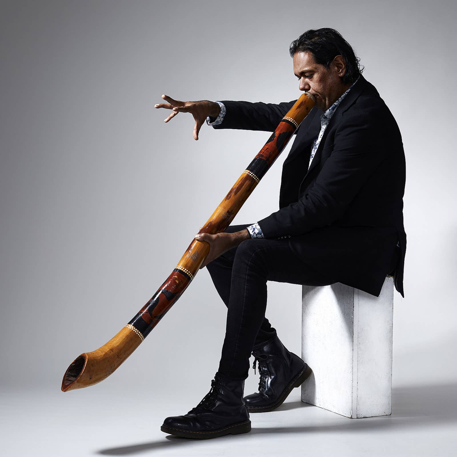 Man in a black suit playing a decorated didgeridoo while seated on a white block against a gray background.