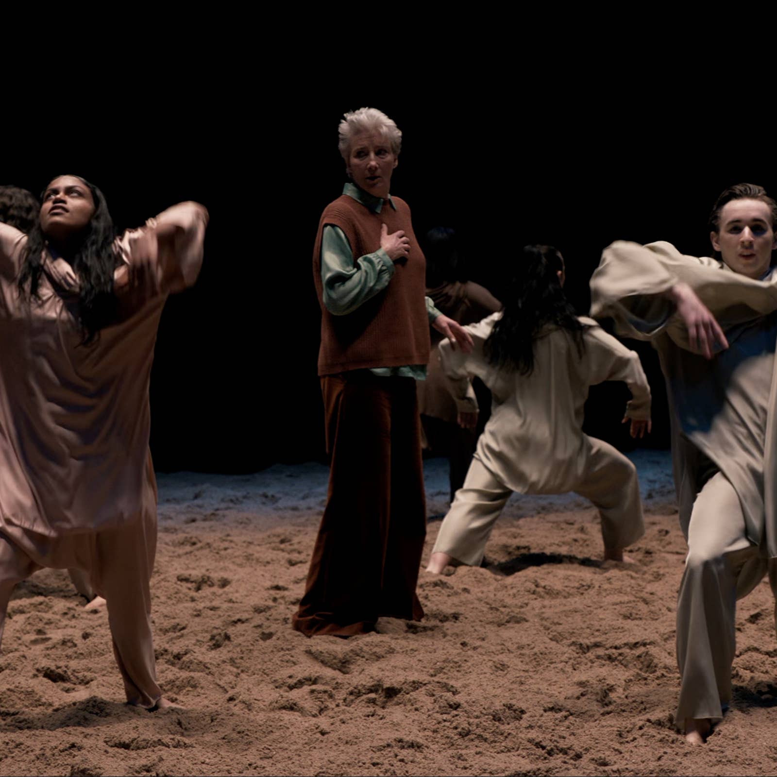 Dancers perform expressively on a sandy stage, wearing loose, neutral-toned clothing, with a dark background.