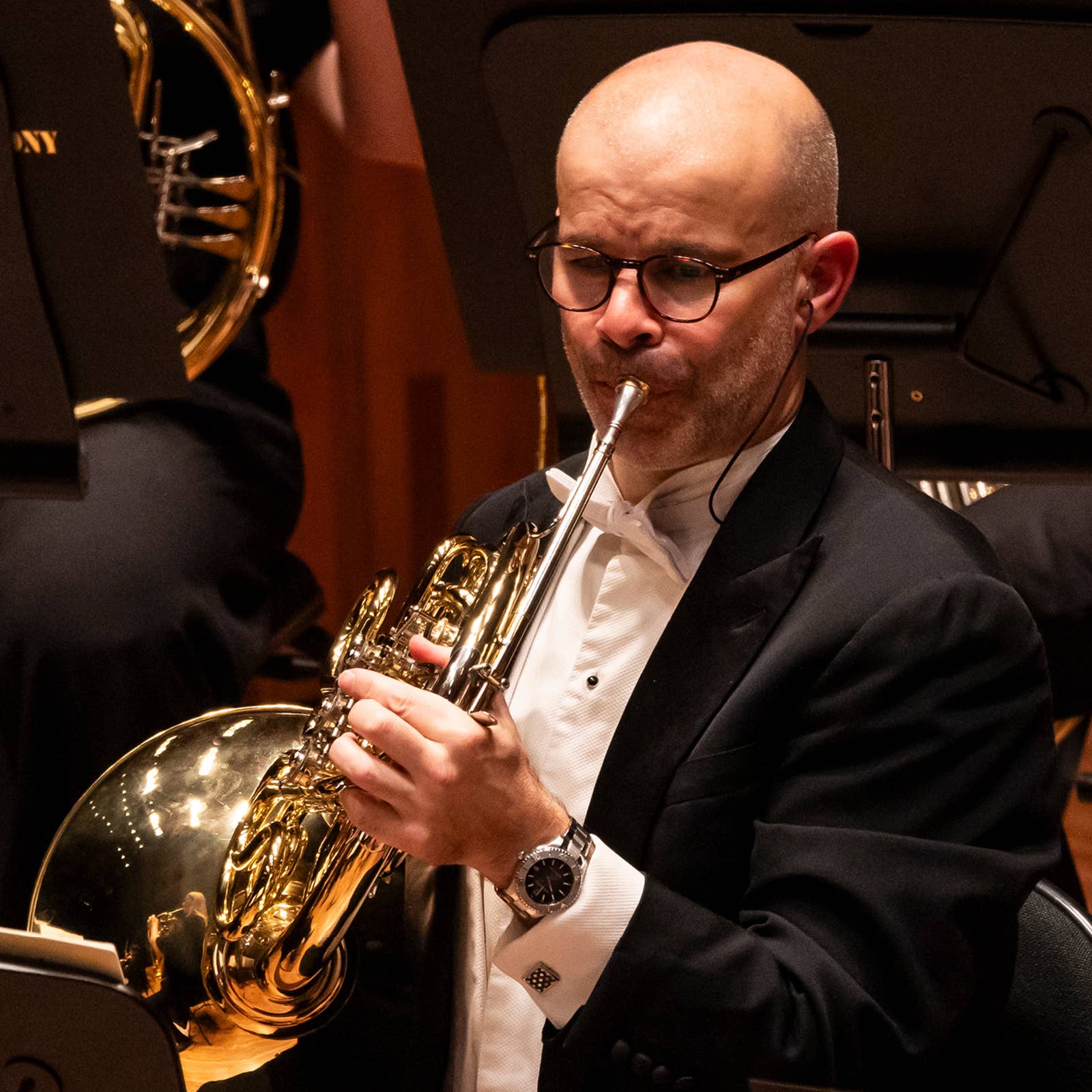 Man in a tuxedo playing a French horn in an orchestra setting.