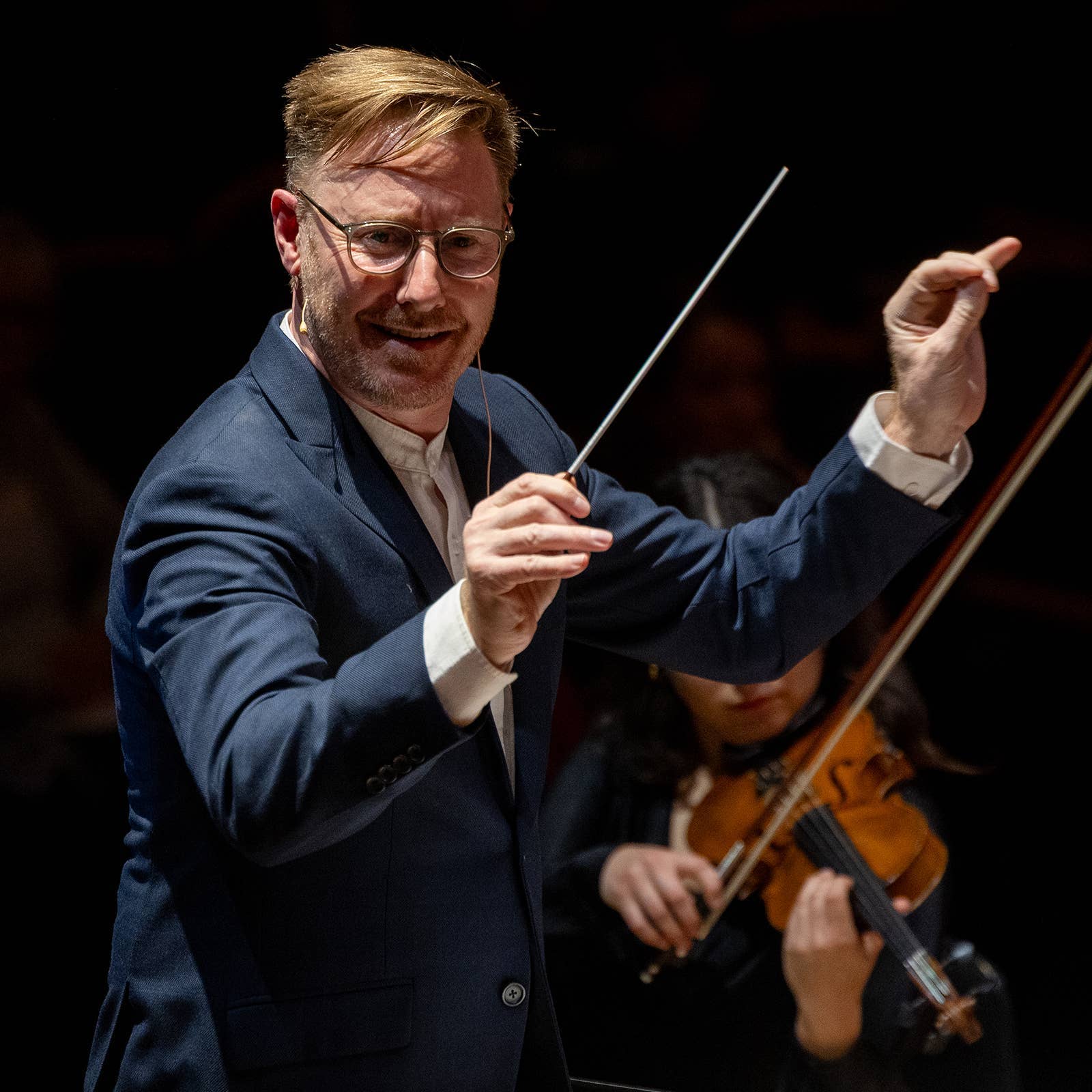 Conductor in a blue suit leading an orchestra, with a violinist in the background.