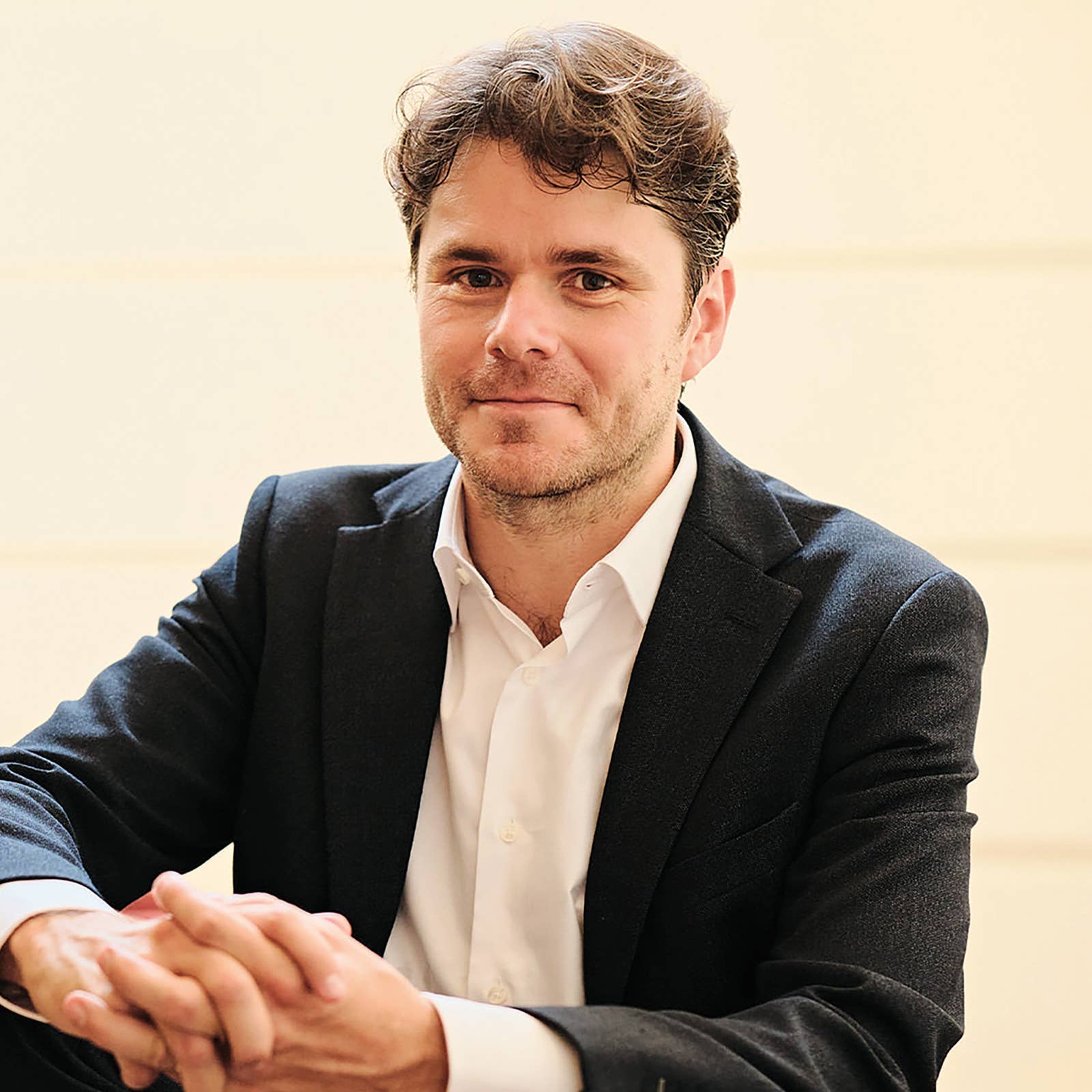 Man in a dark suit and white shirt sitting with hands clasped, smiling slightly against a light background.