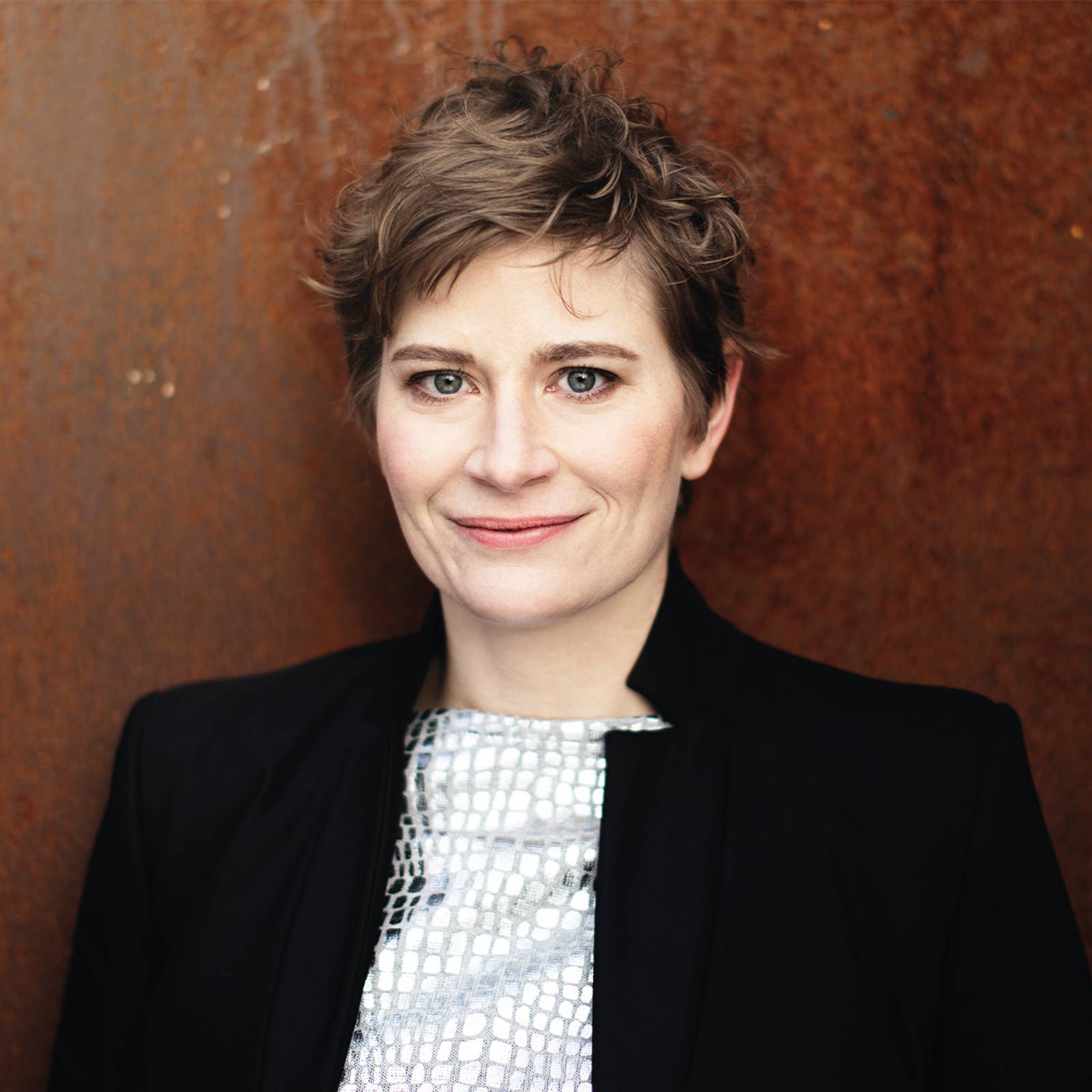 Smiling woman with short hair wearing a black jacket and patterned top, standing against a rust-colored background.