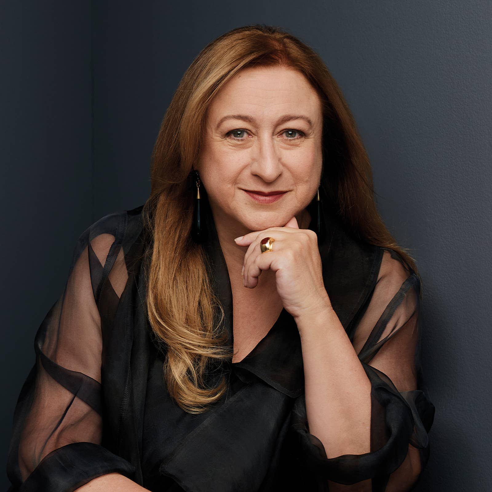 Woman with long hair and sheer black top, resting her chin on her hand, against a dark gray background.