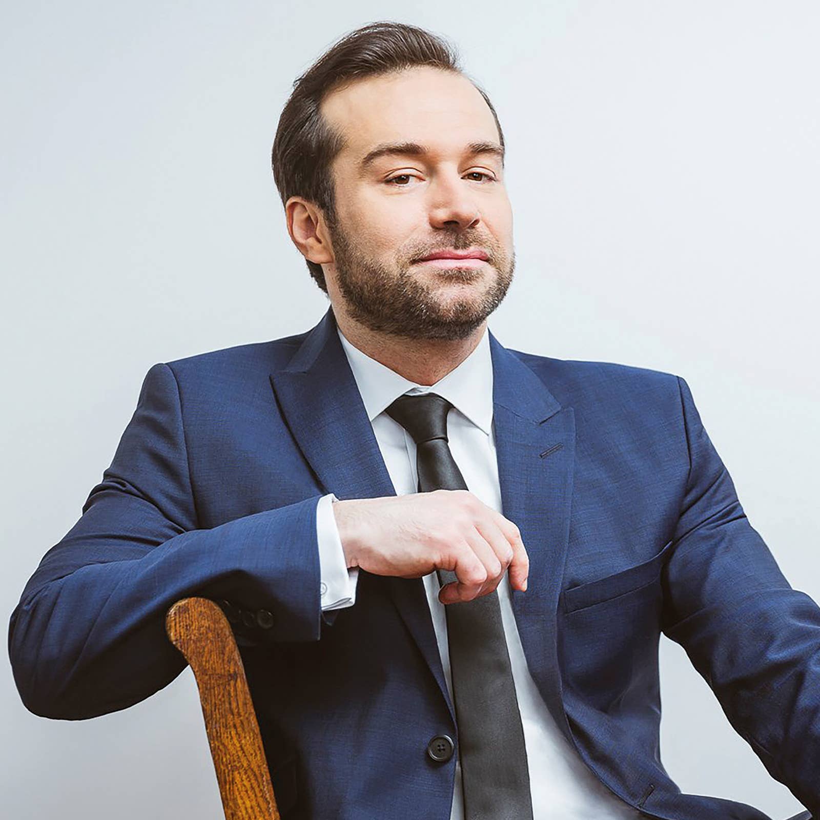 Man in a blue suit and black tie sitting on a wooden chair, looking confidently at the camera against a plain background.
