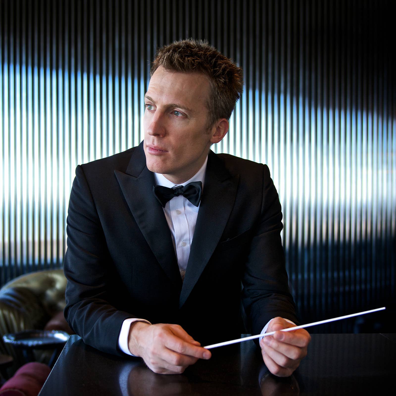 Man in a tuxedo holding a conductor's baton, seated at a table with a striped background.