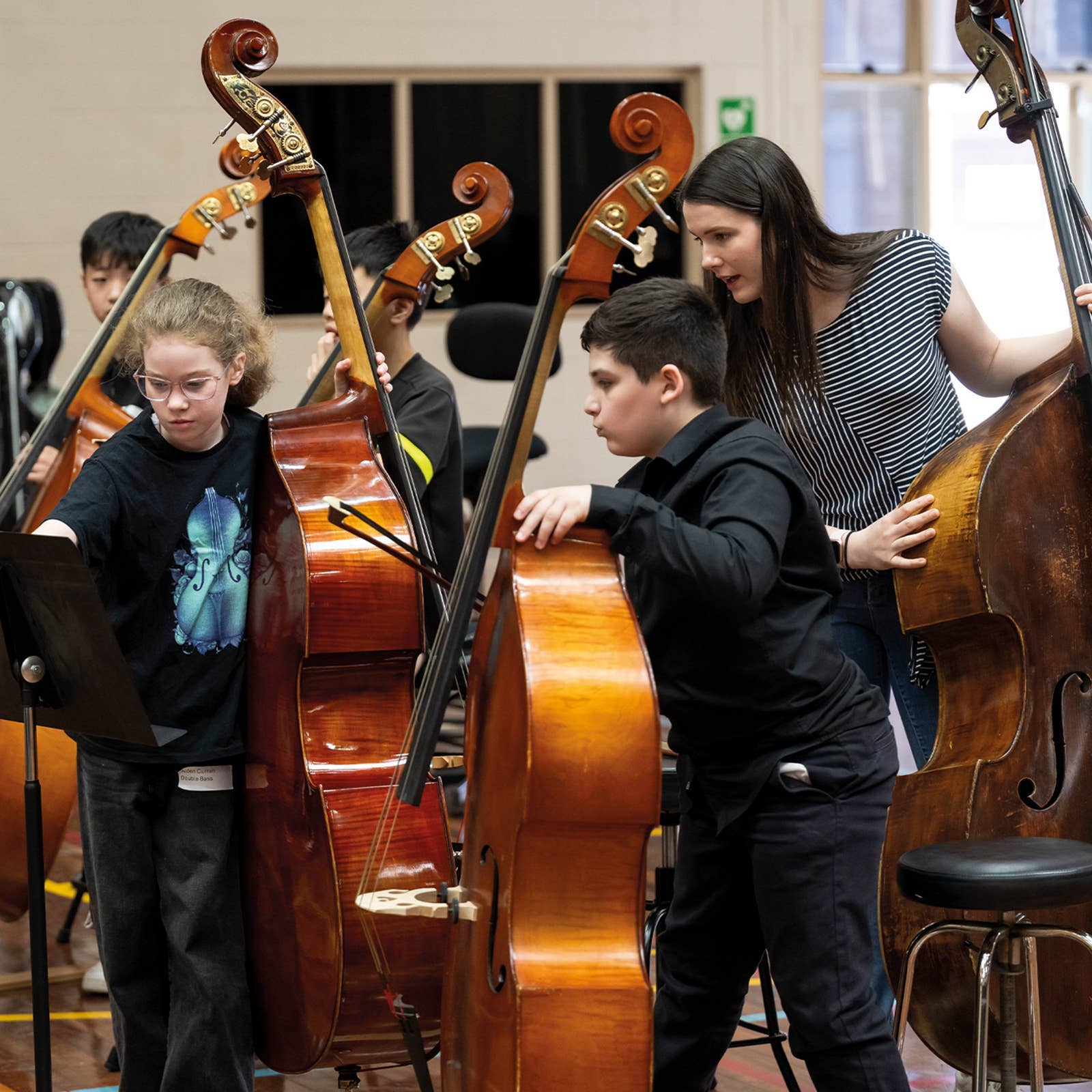 Children playing double basses with guidance from an instructor in a music classroom setting.