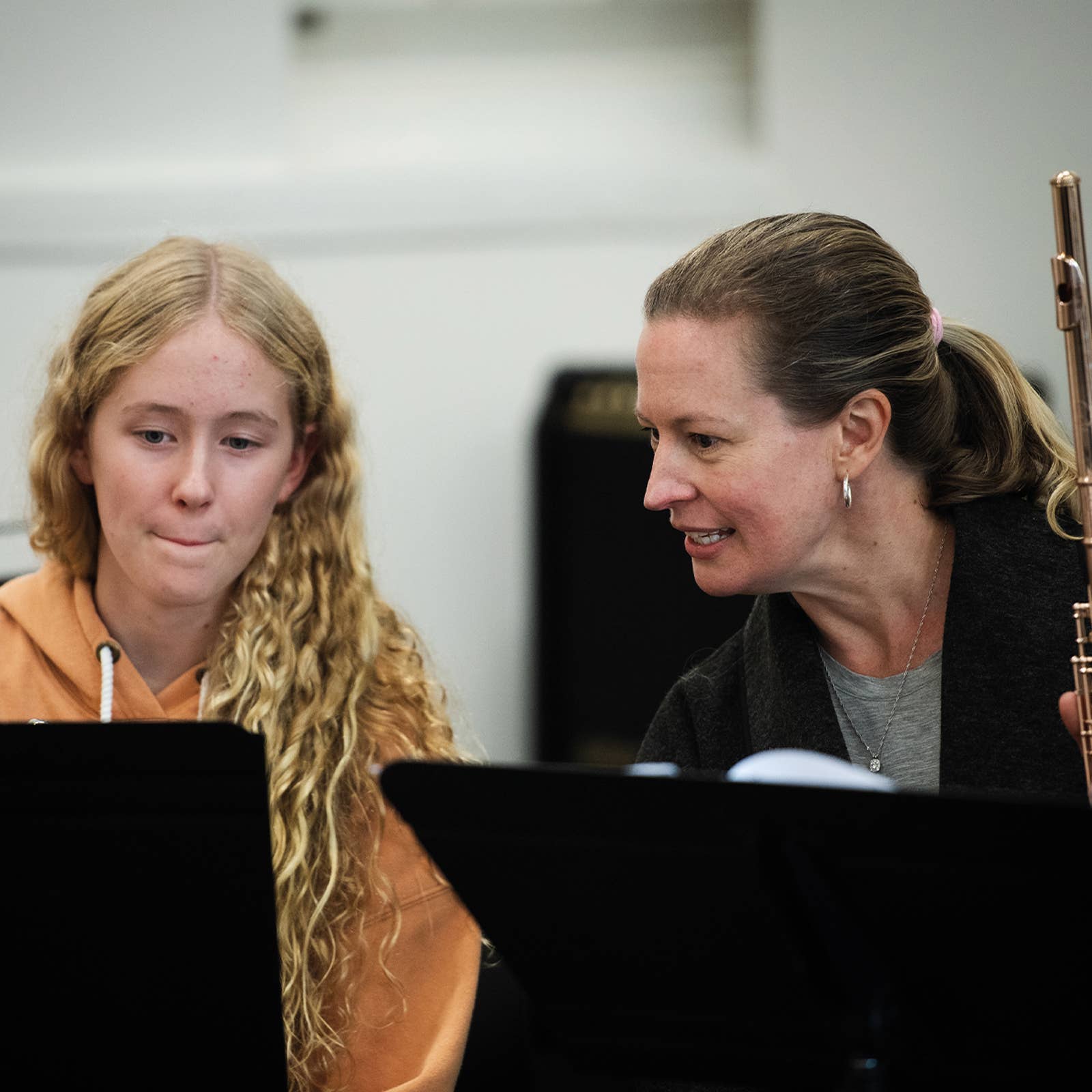 Woman with flute instructs a young girl in a music lesson, both focused on sheet music in a classroom setting.