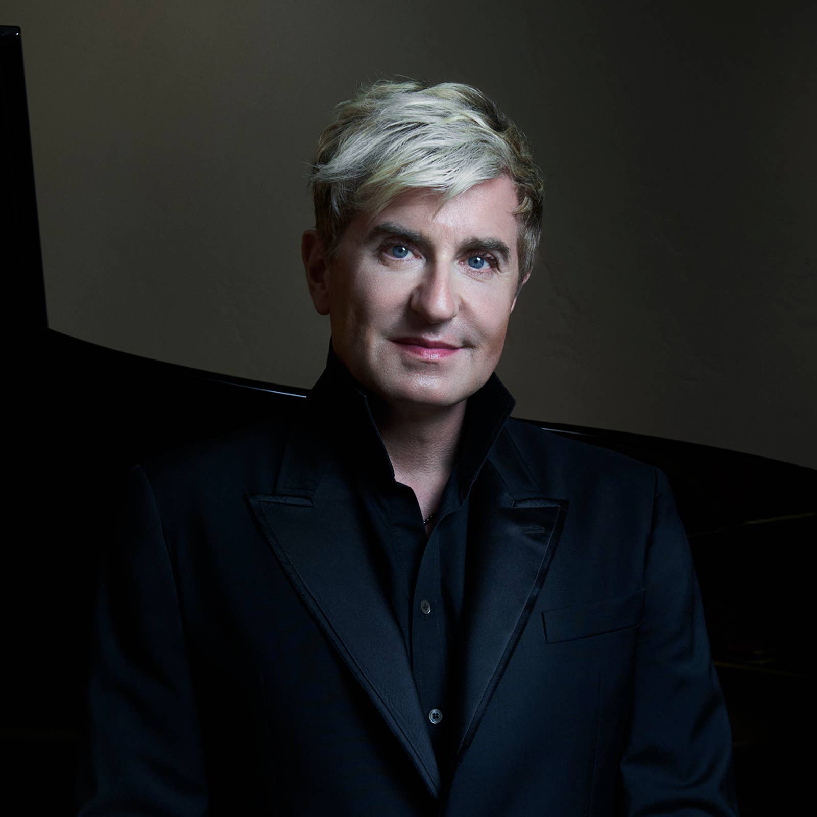 Man with short blond hair wearing a black suit, standing in front of a dark background.