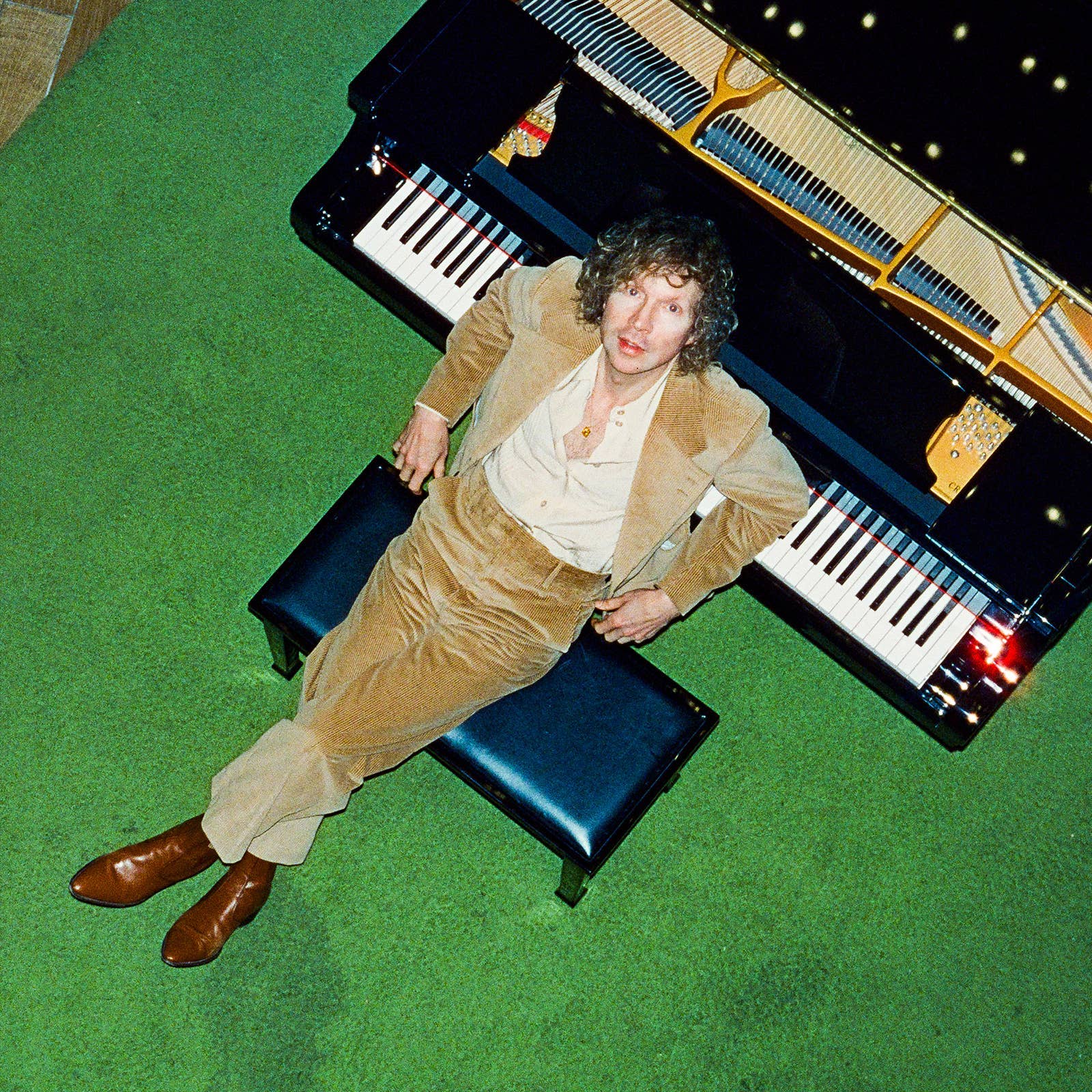 Person in a tan suit sits on a piano bench, viewed from above, with a grand piano and green carpet in the background.