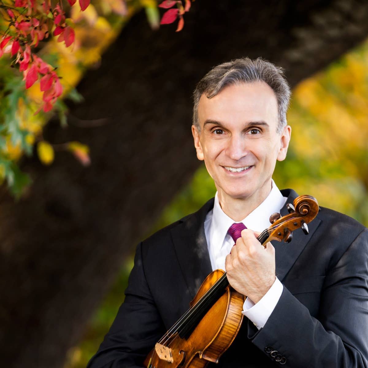 Smiling man in a suit holding a violin outdoors with colorful autumn foliage in soft focus behind him