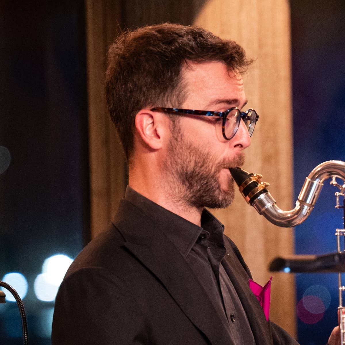 Man in suit playing bass clarinet on stage, wearing glasses, with colorful lights in the background.