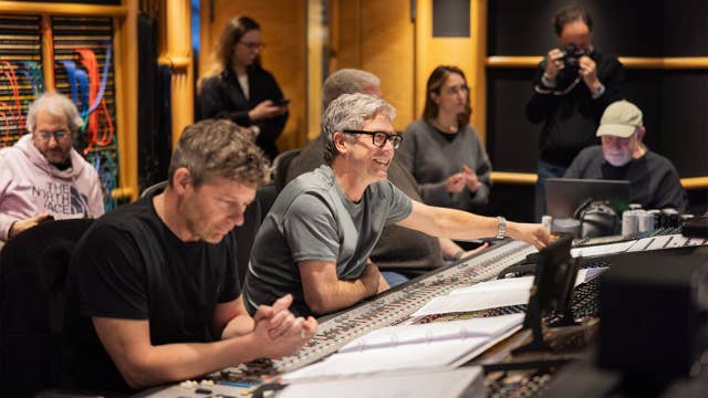 Man with gray hair and glasses smiles while working at a control console in a studio.