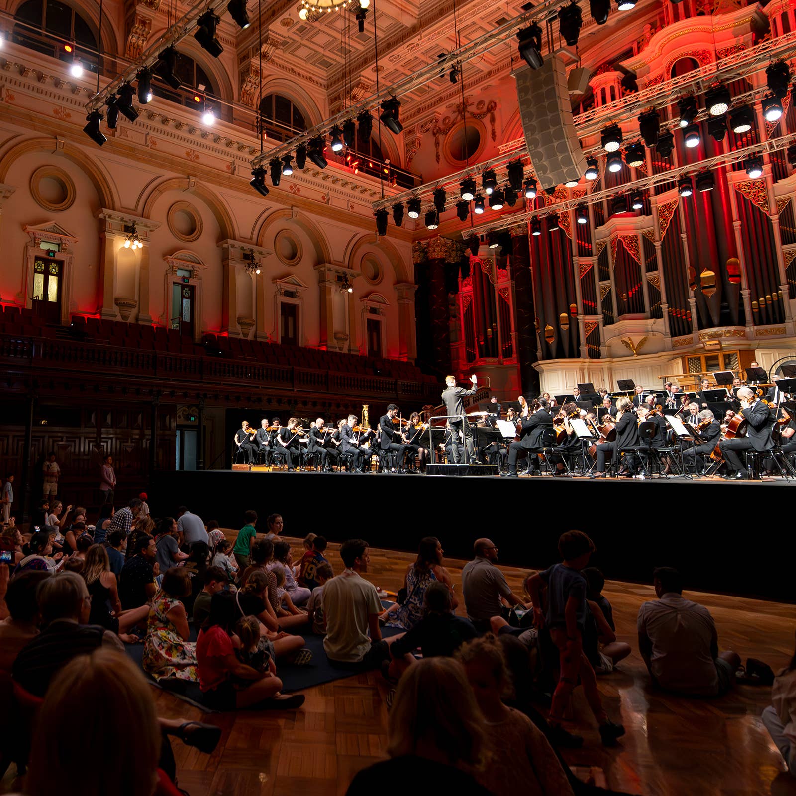 Orchestra performs on stage in a grand concert hall under red lighting, with conductor and seated musicians.