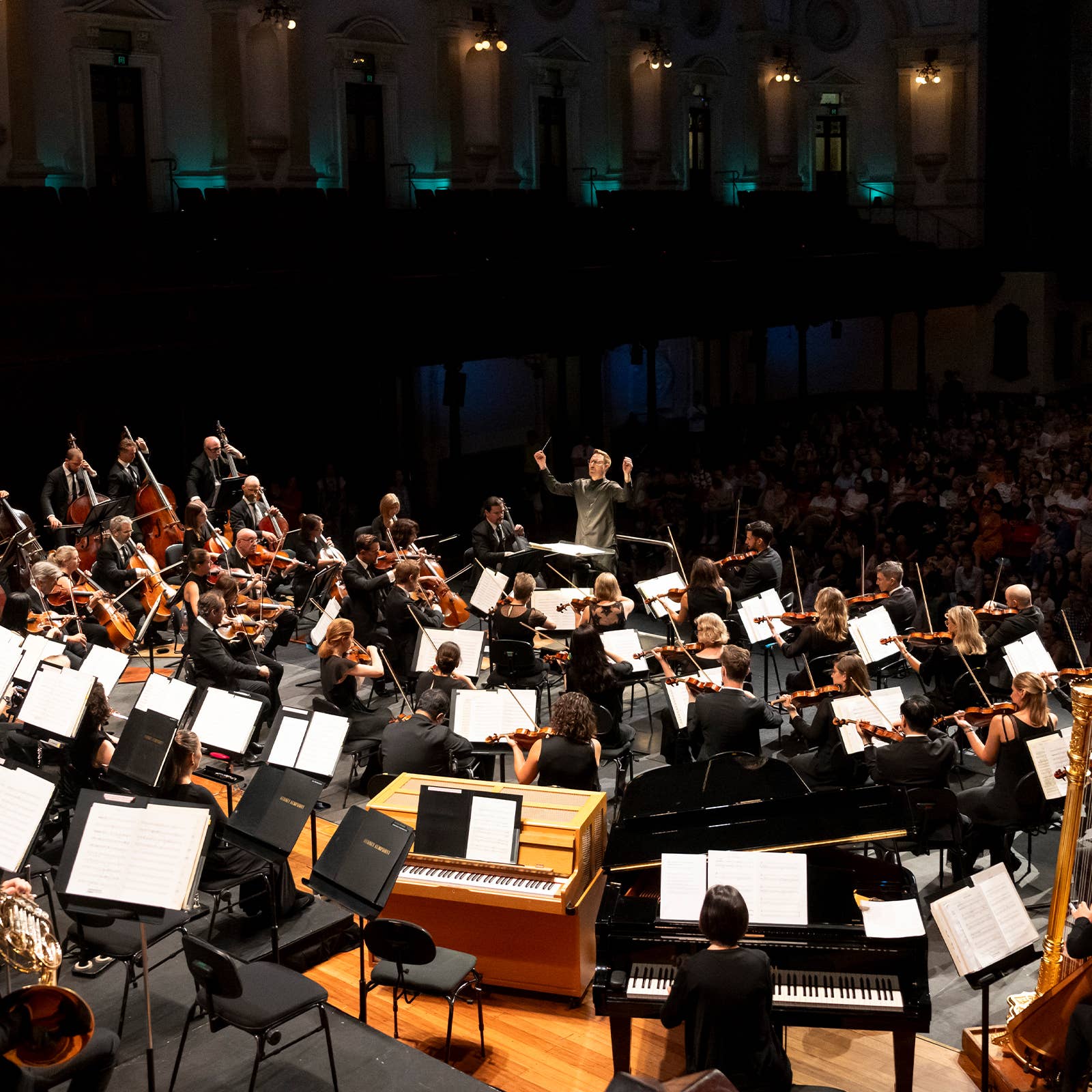 Conductor leads a full orchestra with strings and brass, concert hall seats filled behind in low blue light.