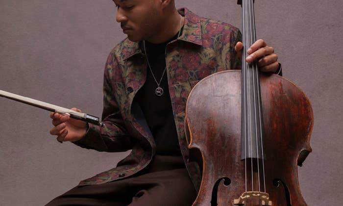Musician seated with a cello, looking down while holding the bow and gripping the fingerboard strings.