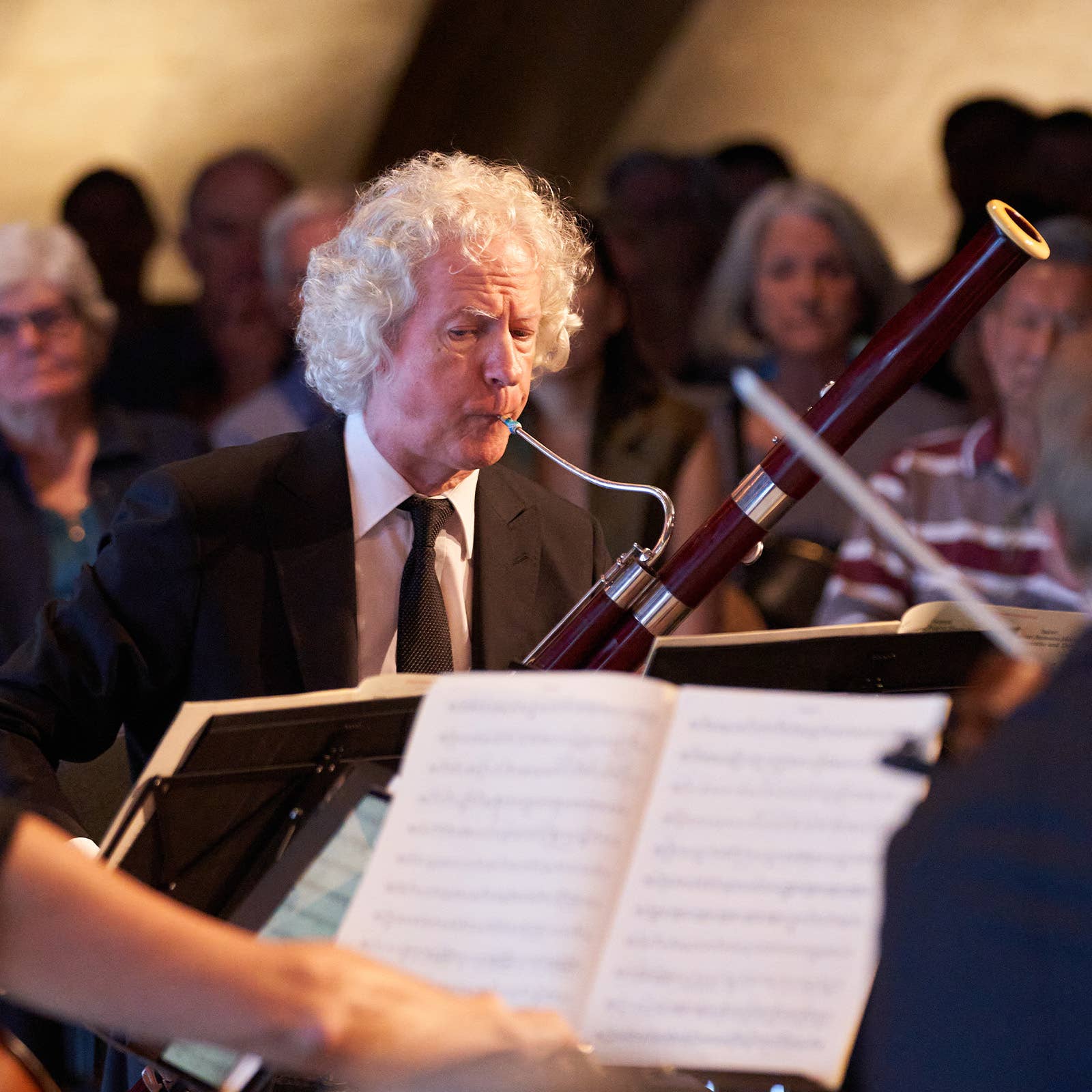 Elderly man with curly white hair performing with a bass clarinet on stage, music stand in foreground, band behind.