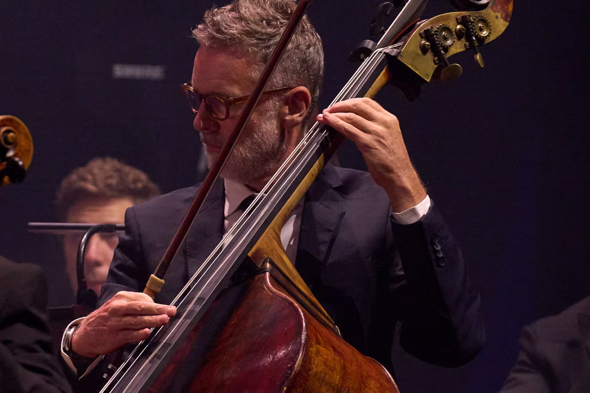 Musician playing an upright double bass on stage, wearing glasses and a dark suit under dim lighting.