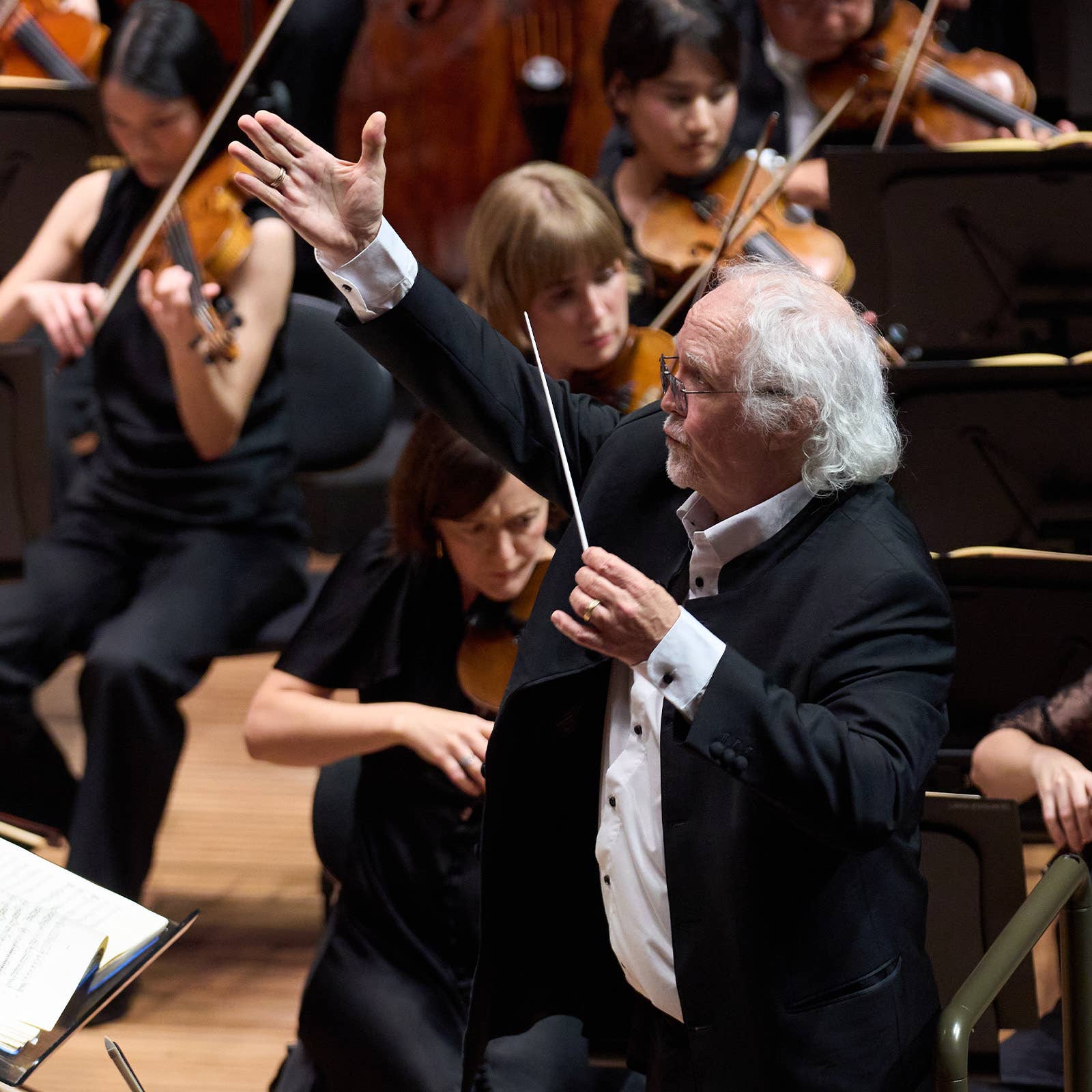 Conductor in black suit raises hand while leading an orchestra with strings in the background