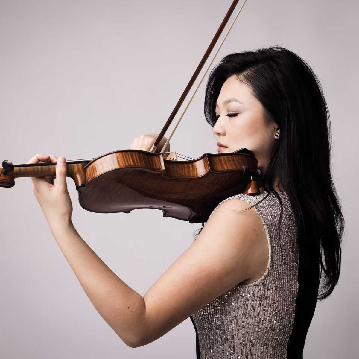 Woman in sequined top playing a violin with eyes closed against a plain light background