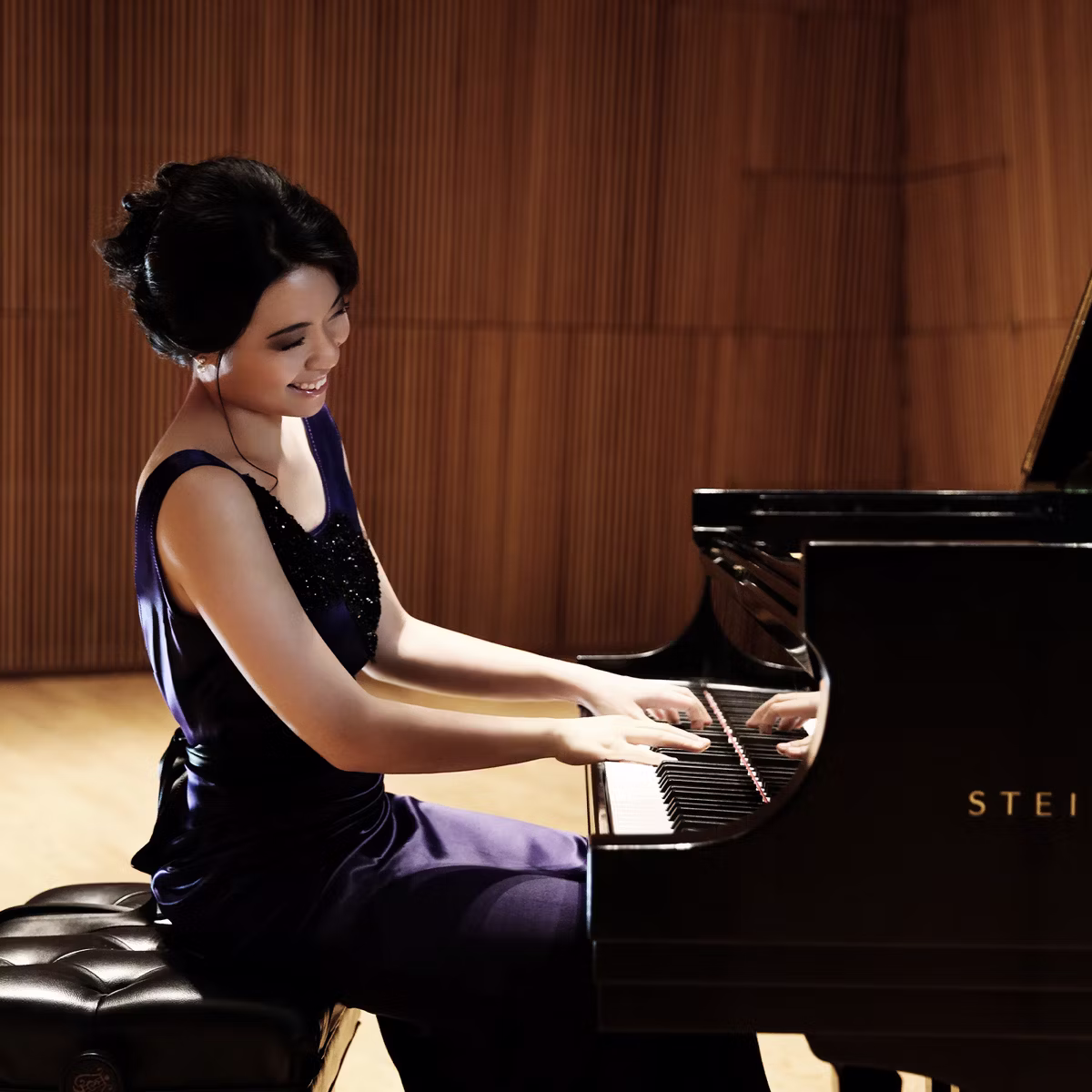 Happy woman smiles while playing a black grand piano in a warmly lit hall