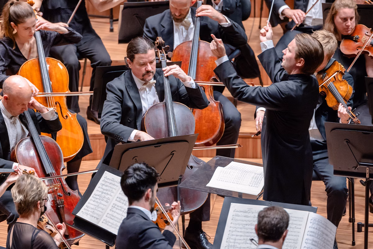 Orchestra musicians in dark suits play strings and double bass while a conductor leads onstage.