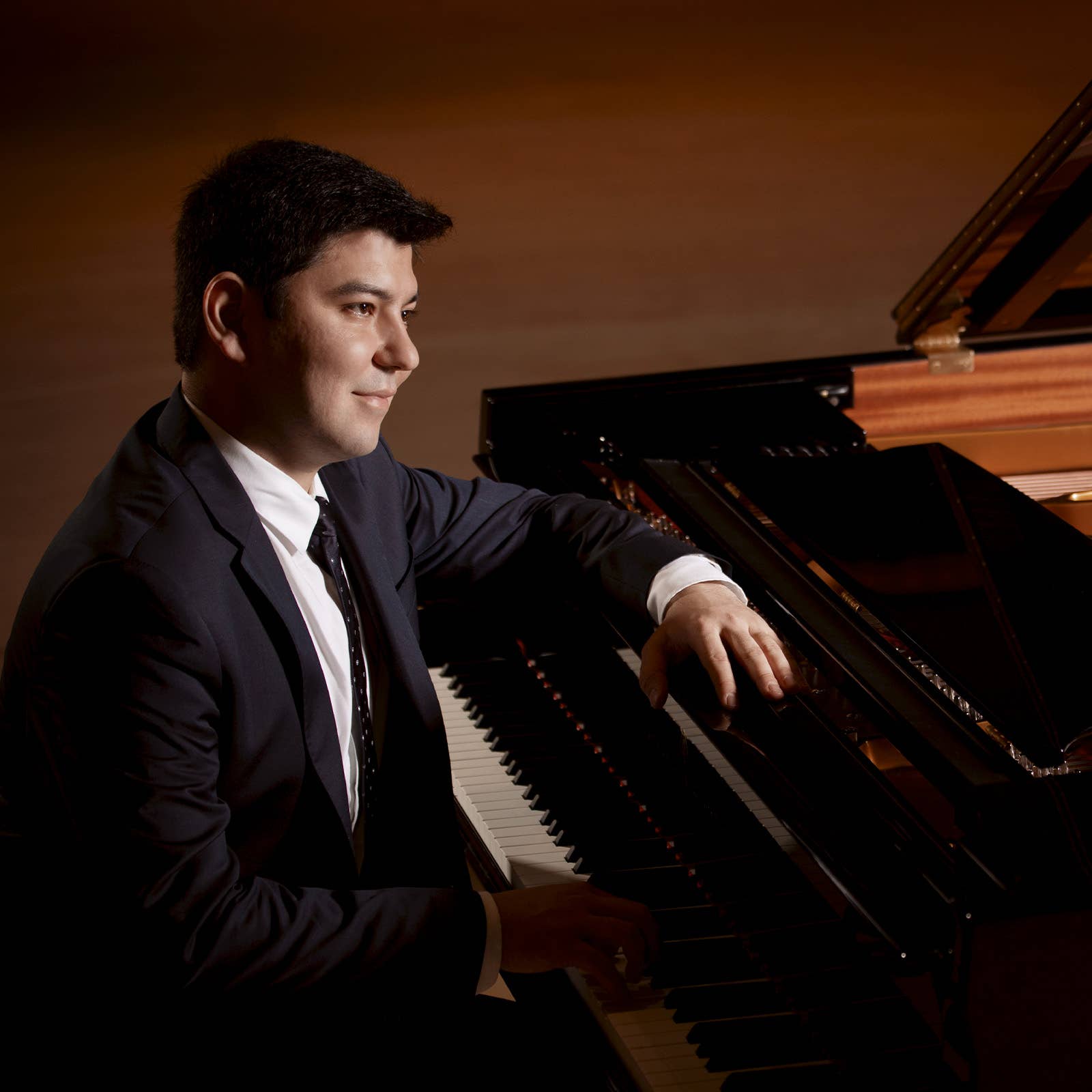 Young man in a suit seated at a black grand piano, smiling under warm studio lighting