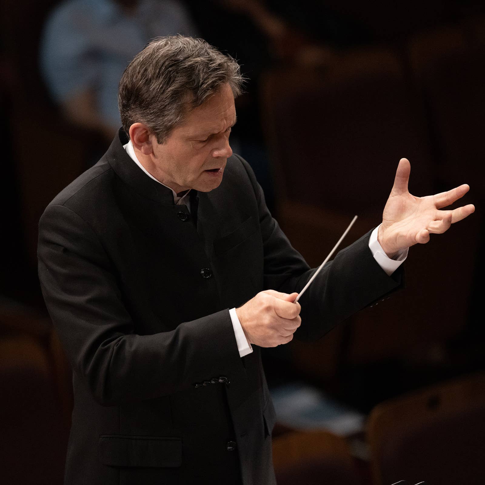 Orchestra conductor in black suit holding a baton and gesturing with an open hand during performance