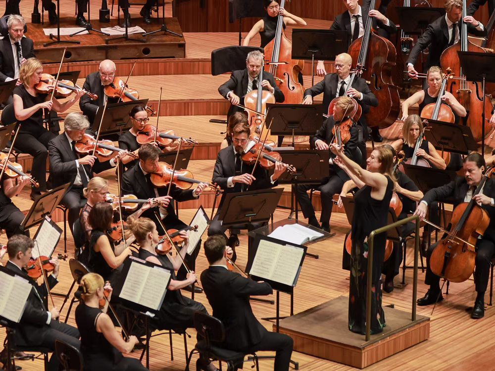 Orchestra performing on stage with violinists and cellists, woodwind and brass players in the background, conductor leading